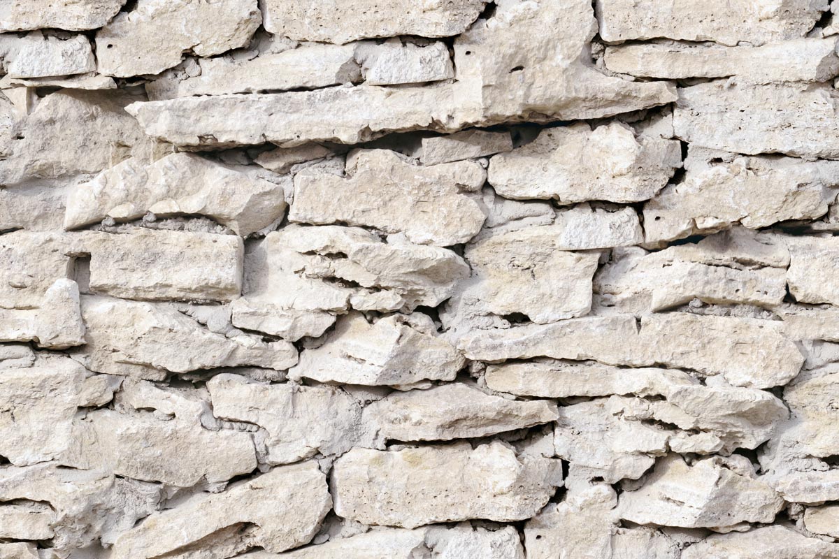 Eine Nahaufnahme einer unebenen Steinmauer aus unregelmäßig geformten, hellen, übereinander gestapelten Steinen mit sichtbaren rauen Strukturen und natürlichen Lücken zwischen den Steinen.