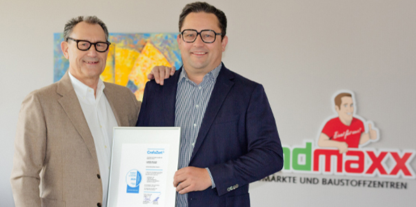 Zwei Männer in Geschäftskleidung stehen lächelnd in einem Raum; einer hält eine gerahmte Urkunde. Im Hintergrund sind ein buntes Kunstwerk und ein Landmaxx-Firmenlogo zu sehen.