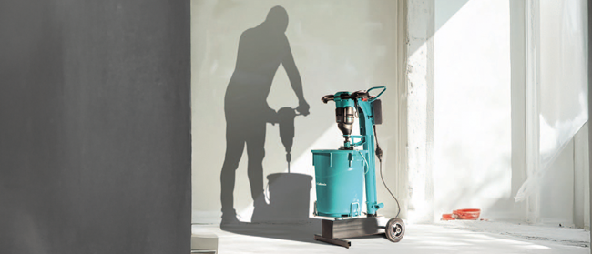 Eine blaugrüne Industriemaschine auf Rädern steht in einem hellen Raum. Ihr Schatten an der Wand erinnert an eine Person, die einen Presslufthammer oder ein Elektrowerkzeug benutzt, obwohl keine Person physisch anwesend ist. Licht fällt von rechts ein.