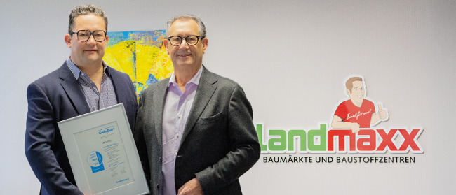 Zwei Männer in Anzügen stehen lächelnd vor einer weißen Wand. Einer hält eine gerahmte Urkunde in der Hand. Dahinter hängt ein buntes Gemälde und ein Logo von landmaxx BAUMÄRKTE UND BAUSTOFFZENTREN mit einer Comicfigur.