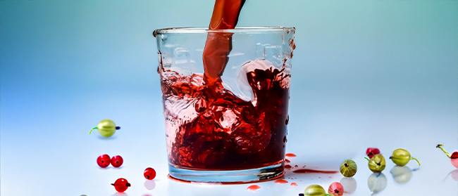 Ein roter Saft wird in ein klares Glas gegossen, umgeben von roten und grünen Beeren auf einer hellblauen Fläche mit blauem Hintergrund.
