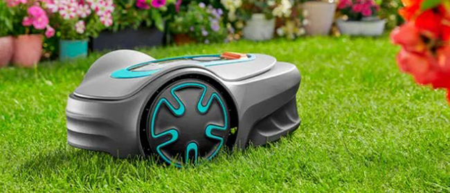 Ein Roboter-Rasenmäher mit schwarzen und blauen Details schneidet Gras in einem Garten, umgeben von blühenden Blumen und grünen Pflanzen im Hintergrund.