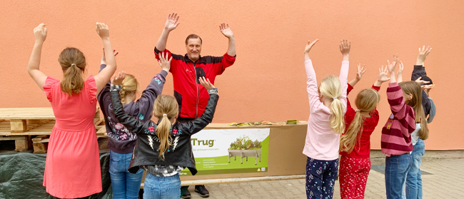 Ein Mann und eine Gruppe Kinder stehen draußen vor einem Tisch mit Pflanzen, heben alle ihre Hände und lächeln begeistert vor einem orangefarbenen Wandhintergrund.