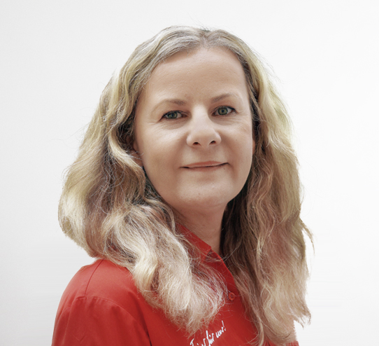 Eine Frau mit langem, gewelltem blondem Haar und heller Haut trägt ein rotes Hemd mit Kragen, steht vor einem schlichten weißen Hintergrund und lächelt leicht in die Kamera.