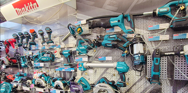 Eine Auslage mit verschiedenen Makita Elektrowerkzeugen, darunter Bohrmaschinen, Sägen und Schleifmaschinen, die auf einer perforierten Wand angeordnet sind, wobei unter jedem Werkzeug Preisschilder und Etiketten sichtbar sind.