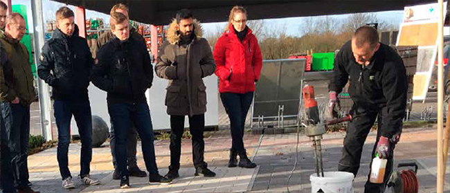 Eine Gruppe von Menschen in Winterjacken steht im Freien und beobachtet einen Mann, der Geräte mit Rohren und einem weißen Eimer auf dem Boden vorführt.