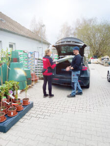 Eine Frau in einer roten Weste übergibt einem Mann, der auf dem Parkplatz eines Gartencenters am offenen Kofferraum eines schwarzen Autos steht, umgeben von Topfpflanzen und Gartenbedarf, einen Sack Erde.