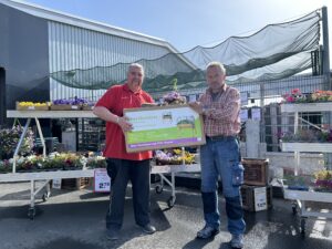 Zwei Männer stehen draußen vor Blumenbeeten in einem Gartencenter. Ein Mann im roten Hemd hält eine Seite eines Mini-Hochbeets, der andere Mann im karierten Hemd die andere Seite.
