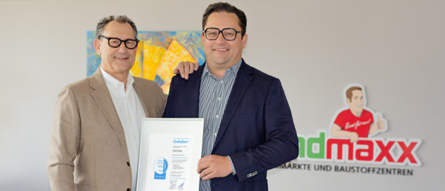 Zwei Männer in Geschäftskleidung stehen lächelnd in einem Raum; einer hält eine gerahmte Urkunde. Hinter ihnen stehen ein farbenfrohes Gemälde und ein Landmaxx-Schild mit einem Cartoon-Logo.