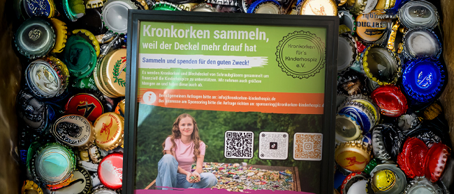 Eine mit bunten Kronkorken gefüllte Schachtel umgibt ein Schild in deutscher Sprache, das für das Sammeln von Kronkorken für wohltätige Zwecke wirbt und ein junges Mädchen zeigt, das auf einer Decke sitzt, sowie QR-Codes für weitere Informationen.